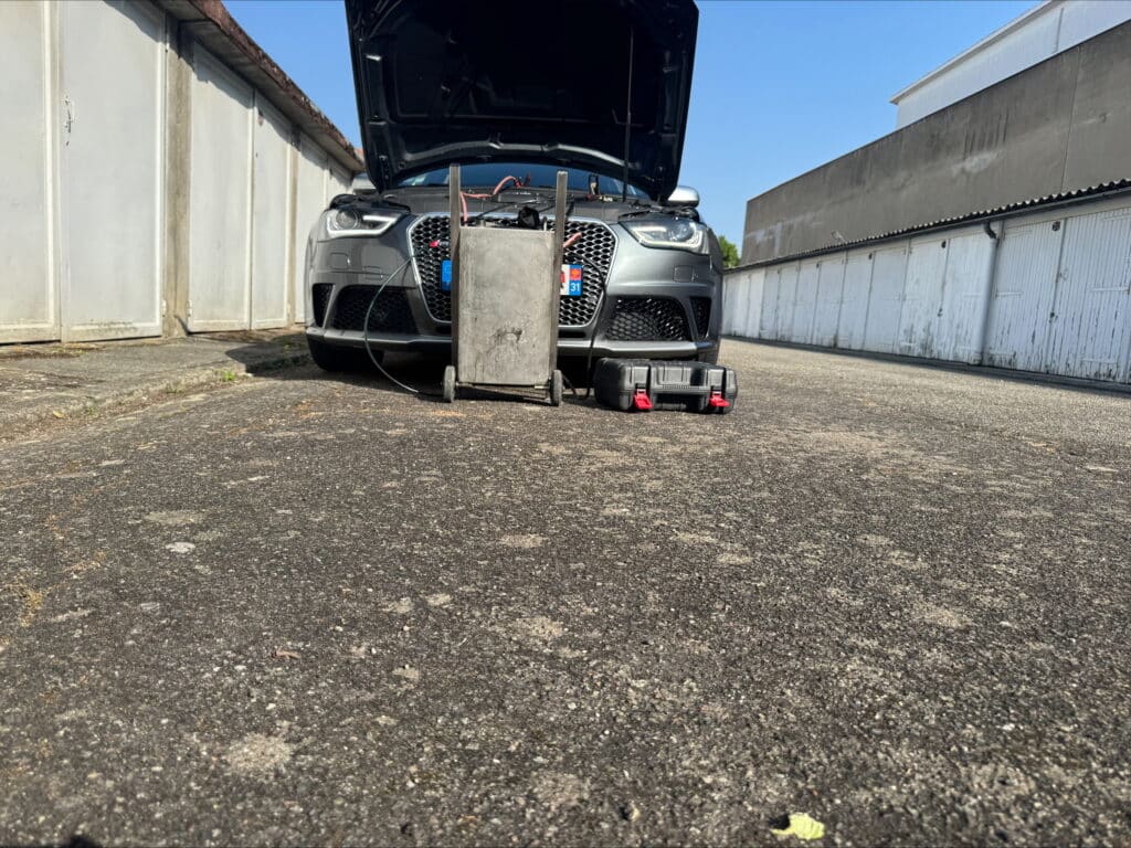 Audi gris foncé en maintenance, capot ouvert, avec un chariot et boîte à outils devant une rangée de garages blancs. Plaque F 31.