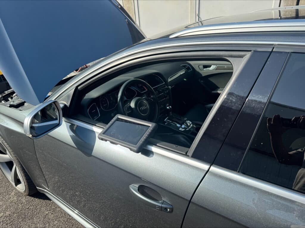Audi RS4 grise avec capot ouvert en plein diagnostic, un outil sur la vitre, et l'intérieur du véhicule visible.