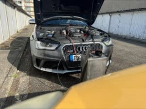 Audi RS4 grise capot ouvert, moteur branché à une machine de nettoyage. Plaques françaises F, garages blancs en arrière-plan ensoleillé.