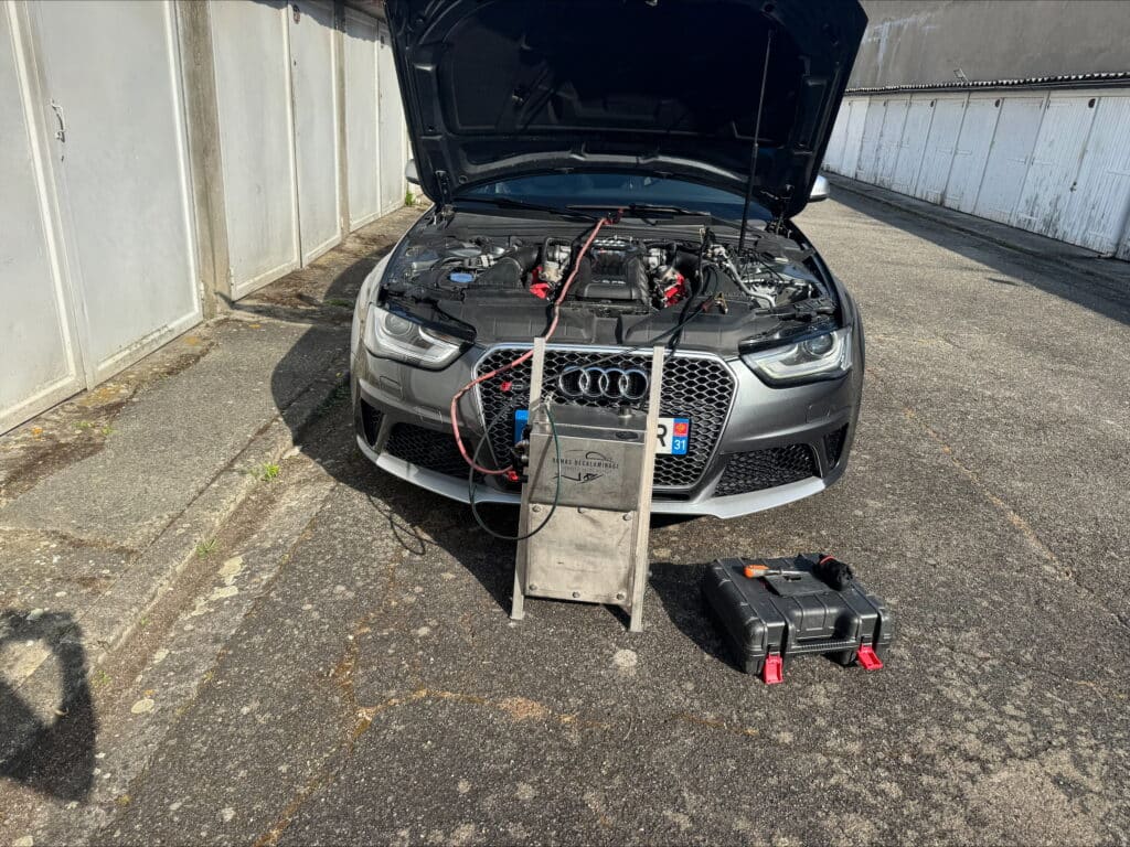 Audi RS grise capot ouvert, équipement de décalaminage "Damas Décalaminage" connecté au moteur. Garages blancs et outils au sol.