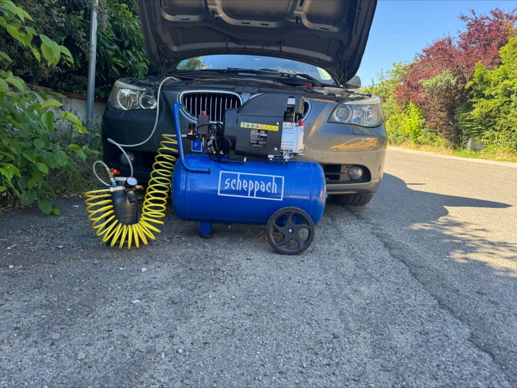Voiture grise avec capot ouvert sur route, un compresseur d'air bleu Scheppach et un tuyau jaune en avant-plan, pour entretien.