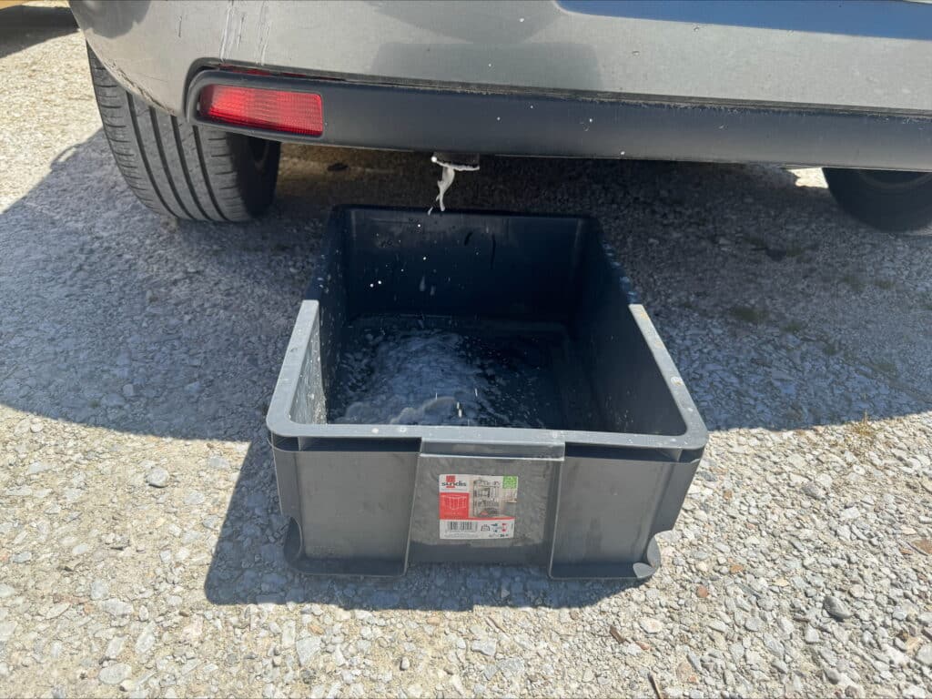 Eau mousseuse s'écoulant d'un drain de voiture grise vers un bac de récupération Sundis gris sur gravier. Entretien véhicule.