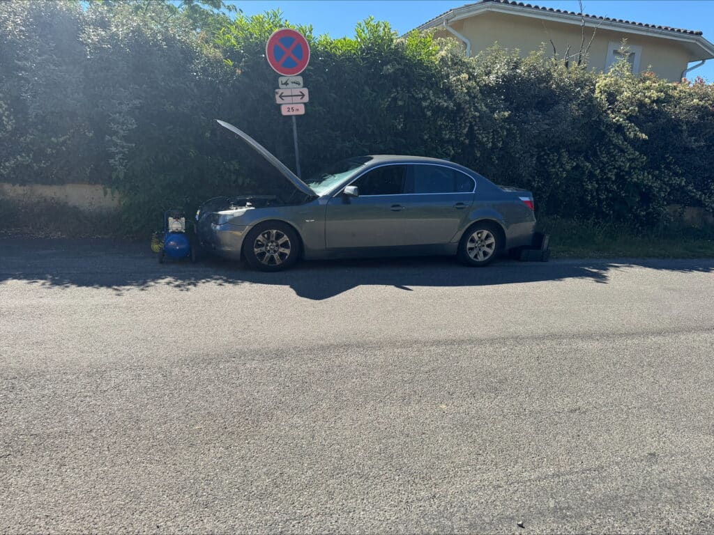 Voiture grise en panne, capot ouvert, sur le bas-côté. Compresseur d'air bleu. Panneau 'Arrêt interdit', 'Dépannage', 'Remorquage 25m'.