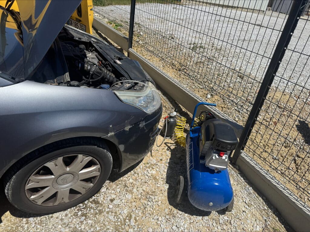 Citroën grise (CN-185-ZG) capot ouvert, moteur exposé. Un compresseur d'air bleu et un réservoir sont connectés pour réparation ou entretien.